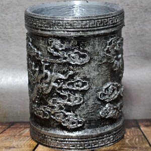 Puede incluir: Un recipiente cilíndrico con acabado plateado y intrincados motivos en relieve de nubes y dragones. El recipiente tiene una superficie texturizada y una banda decorativa cerca de la parte superior. El diseño sugiere un tema de inspiración asiática.