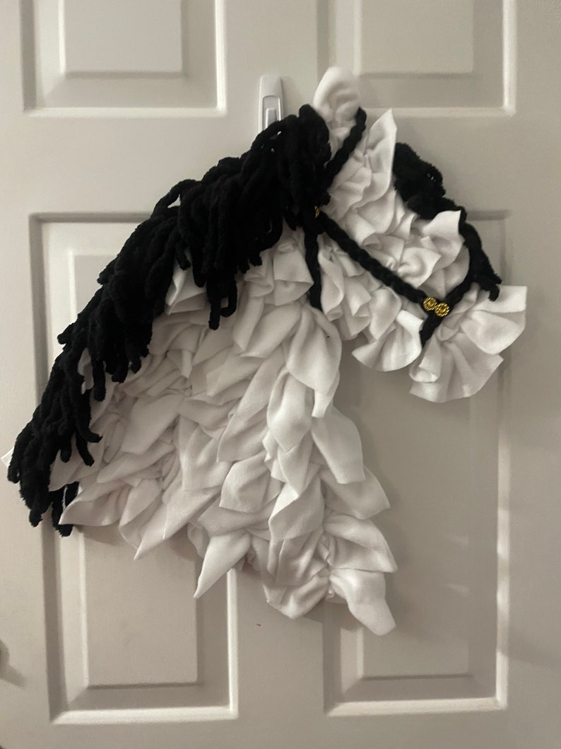 May include: A white and black horse head made of fabric. The horse has a black mane and tail, and a white body with ruffled fabric. The horse is hanging on a white door.