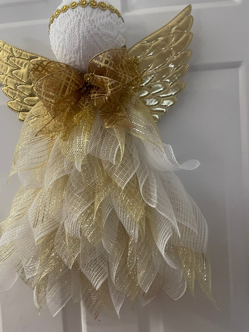 May include: A white angel Christmas decoration with gold wings and a gold bow. The angel is made of mesh and has a white head with a gold trim.
