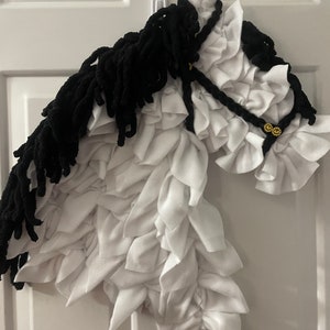 May include: A white and black horse head made of fabric. The horse has a black mane and tail, and a white body with a ruffled texture. The horse is wearing a black bridle with gold accents.