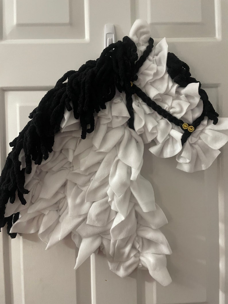 May include: A white and black horse head made of fabric. The horse has a black mane and tail, and a white body with a ruffled texture. The horse is wearing a black bridle with gold accents.