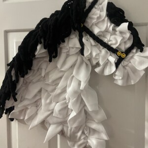 May include: A white and black horse head made of fabric. The horse has a black mane and tail, and the head is made of white fabric with ruffles. The horse head is hanging on a white door.