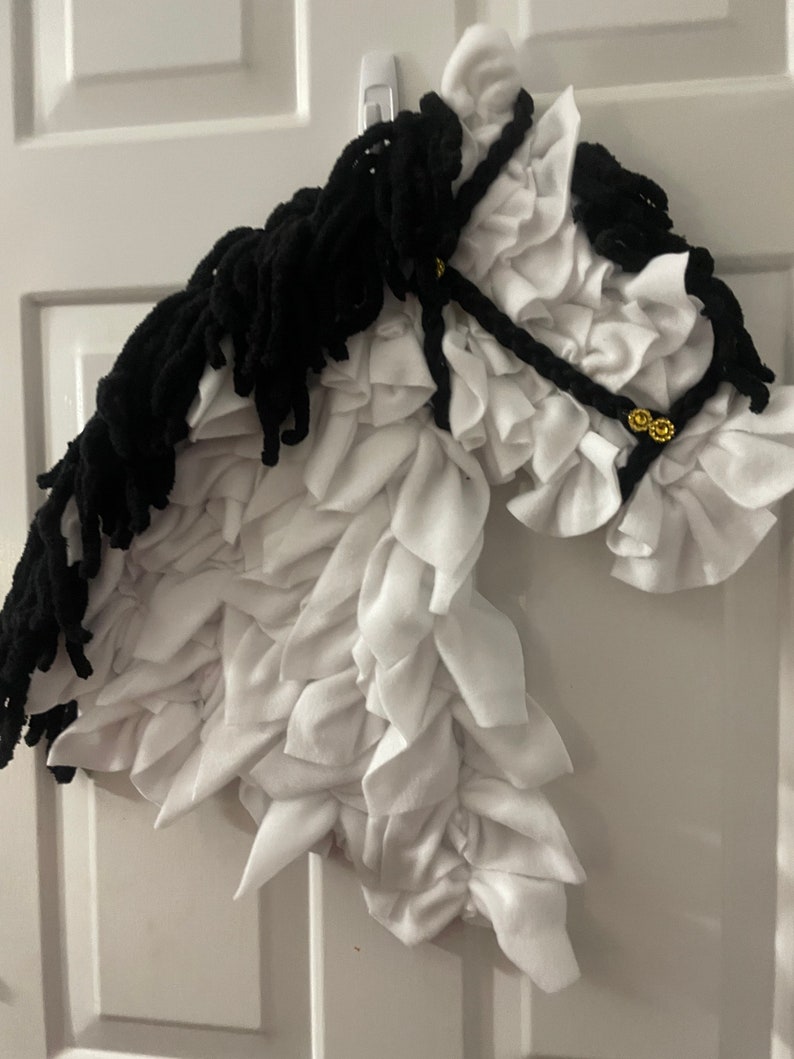 May include: A white and black horse head made of fabric. The horse has a black mane and tail, and the head is made of white fabric with ruffles. The horse head is hanging on a white door.