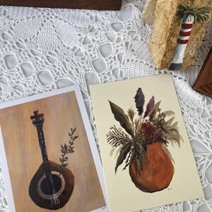 May include: Two framed prints, one with a black and white illustration of a musical instrument, and the other with a watercolor painting of a bouquet of flowers in a terracotta pot.