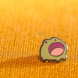 May include: A small, enamel pin featuring a cartoon frog with a wide open mouth. The frog is light green with a pink mouth and a silver outline. The pin is set against a textured, golden-yellow background.