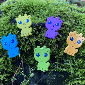 May include: Six colourful cartoon creature charms in various shades of blue, green, yellow, purple, and orange. Each charm has large blue eyes and a sad expression. They are arranged on a bed of green moss, creating a vibrant contrast.