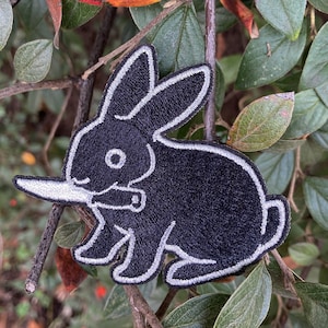 Könnte beinhalten: Schwarzer gestickter Aufnäher mit einem Kaninchen, das ein Messer im Maul hält. Das Kaninchen ist mit weißem Garn umrandet.