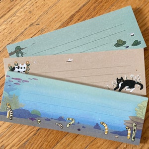 Peut inclure: Ensemble de trois cartes de vœux vierges avec des motifs illustrés. La carte du haut représente une grenouille verte sur un nénuphar, la carte du milieu représente un chat noir et blanc, et la carte du bas représente une scène sous-marine bleue et verte avec des anguilles jaunes.
