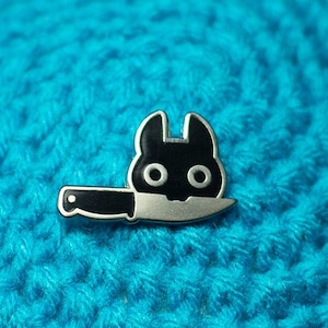 May include: A black and white enamel pin of a cat with a knife through its head. The pin is on a blue crocheted surface.