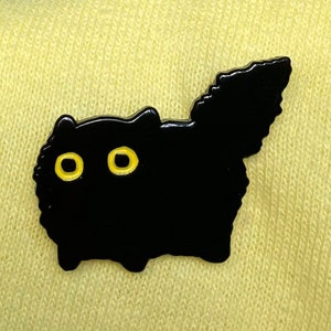 May include: Black enamel pin of a cartoon cat with yellow eyes. The cat is facing forward with its tail curled up behind it.