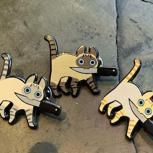 Peut inclure: Trois épingles en émail représentant des chats de dessin animé avec des couteaux dans la bouche. Les chats sont dans différentes poses, l'un tenant le couteau dans sa bouche et les deux autres tenant le couteau dans leurs pattes. Les épingles sont de couleur brun clair, blanc et orange.