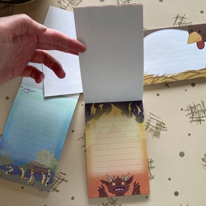 May include: Three blank lined notepads with different illustrations. The first notepad has a blue and green ocean scene with a line of yellow and orange sea creatures. The second notepad has a white background with a black and white illustration of a dragon. The third notepad has a brown and white illustration of a chicken.