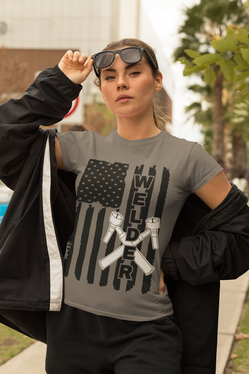 Puede incluir: Una camiseta gris con un dise&ntilde;o de bandera estadounidense en blanco y negro. El texto "WELDER" est&aacute; impreso verticalmente en el lado derecho de la bandera, con dos sopletes de soldadura cruzados.