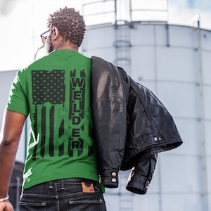 Puede incluir: Camiseta verde con un dise&ntilde;o de bandera estadounidense en blanco y negro y la palabra "WELDER" con herramientas de soldadura cruzadas.
