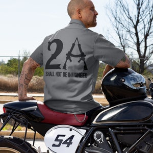 May include: A man wearing a gray short-sleeved shirt with the text "2A SHALL NOT BE INFRINGED" and a graphic of two rifles. He is sitting on a black motorcycle with a red seat and a white number plate that says "54".