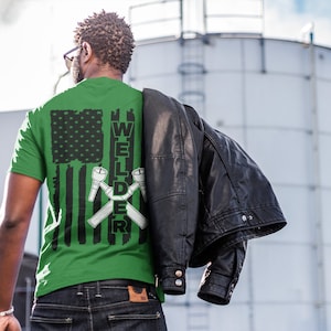 Puede incluir: Camiseta verde con un dise&ntilde;o de bandera estadounidense en blanco y negro y la palabra "WELDER" con antorchas de soldadura cruzadas.