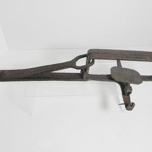 May include: A rusty, antique metal trap. The trap features a rectangular frame, a hinged lever, and a circular loop. The metal has a weathered appearance, suggesting age and use. The trap is designed to catch animals.