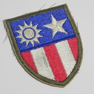 May include: A green, red, white, and blue embroidered patch with a shield shape. The shield has a white star and sun on a blue background, and red and white stripes on the bottom.