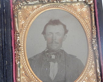 May include: A black and white tintype photograph of a man in a suit and bow tie. The photograph is framed in a gold-colored frame with ornate details.