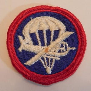 May include: A circular patch with a red border, blue center, and embroidered design. The design features a white parachute, a white airplane, and a propeller. The patch is likely a military or aviation emblem.