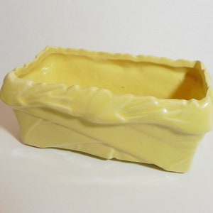 May include: A rectangular, pale yellow ceramic planter. The planter has a textured, folded design around the sides and a scalloped edge. The interior is smooth and the planter is empty.