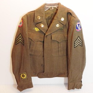 May include: A vintage olive drab military jacket with gold buttons, patches, and insignia. The jacket features two front pockets, epaulets, and rank chevrons on the sleeves. The jacket is hanging on a white hanger against a white background.