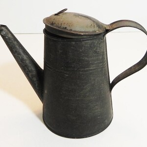 May include: A vintage metal watering can with a long spout and a curved handle. The can is dark gray with a slightly weathered appearance, and a hinged lid. The design is simple and functional, suggesting a rustic or antique aesthetic.
