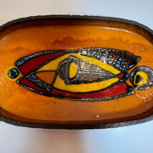 May include: An oval ceramic dish with a dark brown rim and a glossy orange glaze. The dish features a stylized abstract design in black, red, yellow, and blue.
