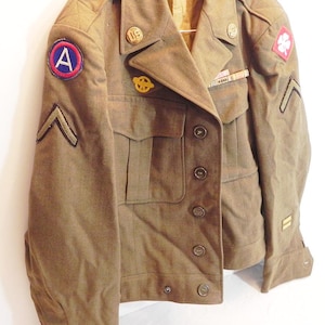 May include: A vintage olive drab military jacket with multiple pockets, buttons, and patches. The jacket features a patch with a red and blue "A" on the left sleeve, a clover patch, and rank chevrons. It has a classic, tailored design.