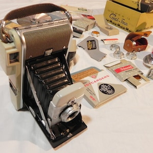 May include: A vintage Polaroid Land Camera with a brown leather strap, a flash attachment, and a variety of accessories, including a Polaroid filter, a lens cap, and a box of Polaroid film.