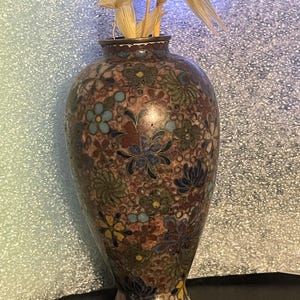 May include: A decorative vase with a floral pattern in shades of brown, blue, green, and yellow. The vase has a narrow neck and a wider body. Dried plants are placed inside the vase. The vase is made of metal.