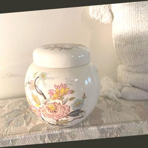 May include: A white ceramic jar with a lid, decorated with a floral pattern in pink, yellow, and blue. The jar is round and rests on a patterned surface. The lid also has a floral design.