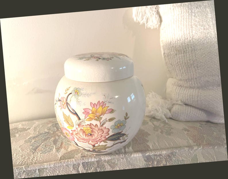 May include: A white ceramic jar with a lid, decorated with a floral pattern in pink, yellow, and blue. The jar is round and rests on a patterned surface. The lid also has a floral design.