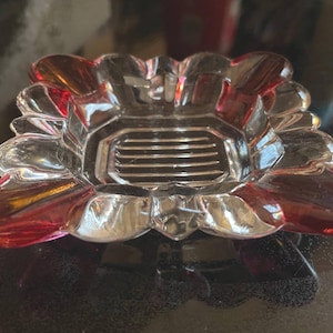 May include: A clear glass dish with a flower-like design. The dish has a central square area with horizontal lines and petal-shaped edges with red accents. The glass reflects light, creating highlights and shadows.