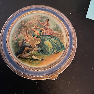 Op de afbeelding: Een ronde, vintage compact met een gedetailleerde geschilderde scène van figuren in een tuin. De compact heeft een blauwe en gouden rand, en het kunstwerk toont mensen in kleding uit die tijd. Een verzamelobject.