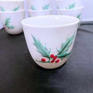 Puede incluir: Tazas de cerámica blanca con un diseño de acebo pintado a mano, con hojas verdes y bayas rojas. Las tazas son cilíndricas y parecen ser parte de un juego, perfectas para reuniones festivas o decoración de temporada.