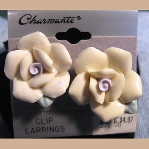 May include: Cream-coloured flower clip earrings with a light purple centre and green leaves. The earrings are on a grey card with the word "Charmante" and "CLIP EARRINGS" printed on it.