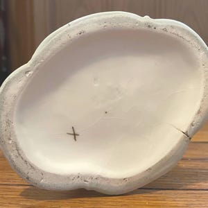 May include: Off-white ceramic decorative piece with a scalloped edge. The interior is smooth and white, with a small cross mark and a crack. The exterior edge is a light green color.