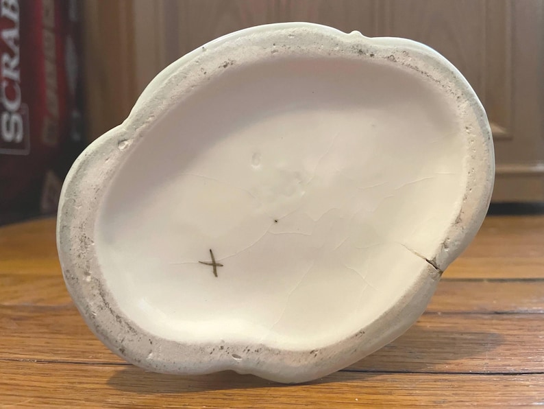 May include: Off-white ceramic decorative piece with a scalloped edge. The interior is smooth and white, with a small cross mark and a crack. The exterior edge is a light green color.