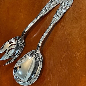 Pode incluir: Um conjunto de servir salada de prata com um design ornamentado. O conjunto inclui uma colher de servir ranhurada e um garfo.