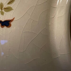 May include: Close-up of a cream ceramic vase with a cracked glaze. The vase is decorated with a blue and orange butterfly and green and brown leaves. The surface has a network of fine cracks.
