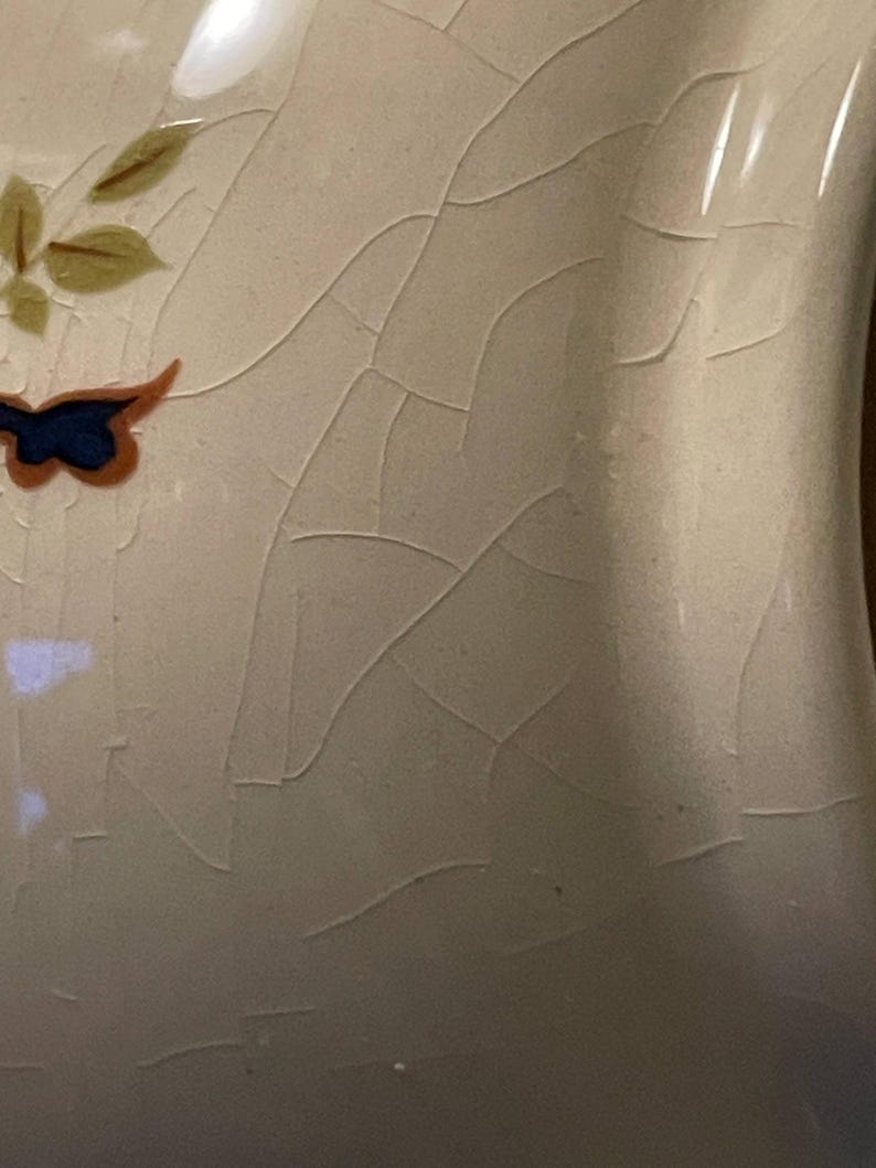 May include: Close-up of a cream ceramic vase with a cracked glaze. The vase is decorated with a blue and orange butterfly and green and brown leaves. The surface has a network of fine cracks.