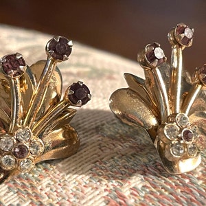 以下が含まれることがあります： 花柄のゴールドトーンのクリップ式イヤリングのペア。イヤリングは、各花の真ん中にパープルのラインストーンのクラスターが特徴です。