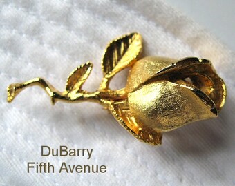 Könnte beinhalten: Eine goldfarbene Rosenbrosche mit strukturierter Oberfläche. Die Rose ist eine einzelne Blüte mit Stiel und Blättern. Die Brosche ist mit "DuBarry Fifth Avenue" signiert.