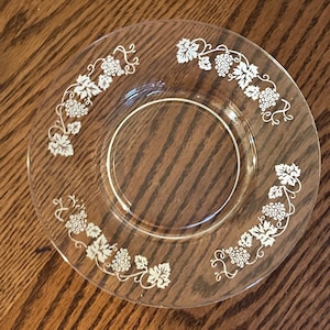 Pode incluir: Um prato de sobremesa de vidro transparente com um desenho gravado branco de videiras e uvas ao redor da borda.