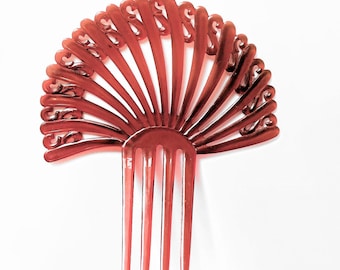 May include: A reddish-brown hair comb with a fan-like design. The comb features a series of vertical tines at the base, with a decorative, curved pattern at the top. The comb is made of a translucent material and is set against a white background.