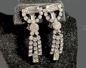 May include: A pair of silver tone dangle earrings with clear rhinestone accents. The earrings feature a large, round rhinestone in the center of each dangle, surrounded by smaller rhinestones.