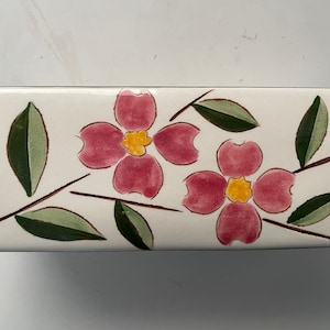 May include: A white ceramic rectangular dish with a floral design. The dish features two pink flowers with yellow centers and green leaves.