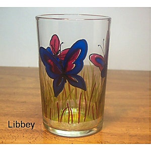 May include: A clear glass tumbler with a painted design of blue and pink butterflies in a field of green and red grass.