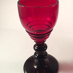 May include: A red glass goblet with a dark red stem and base. The goblet has a rounded bowl and a thick, textured stem.